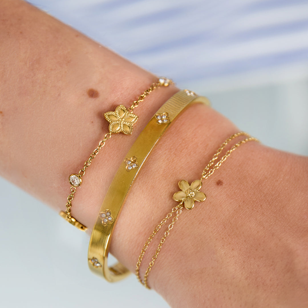 Close-up van een goudkleurige dubbele minimalistische armband met een bloem hanger gedragen om een pols