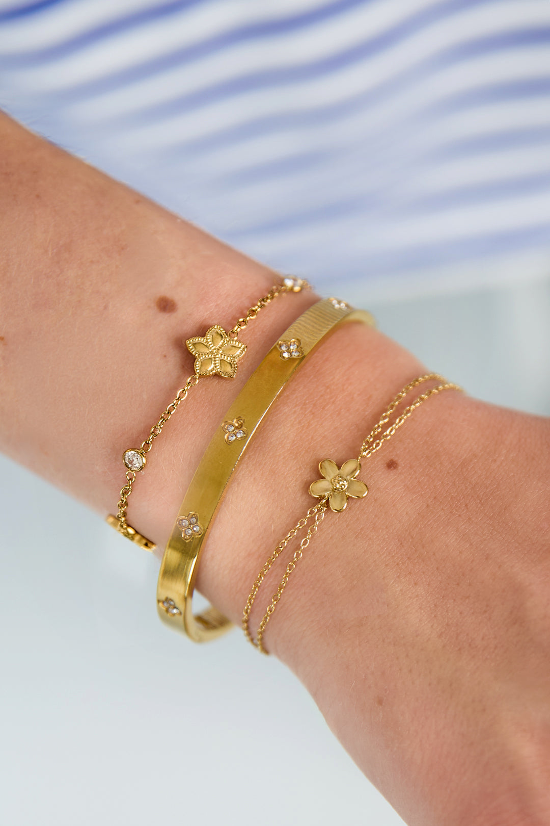 Close-up van een goudkleurige dubbele minimalistische armband met een bloem hanger gedragen om een pols
